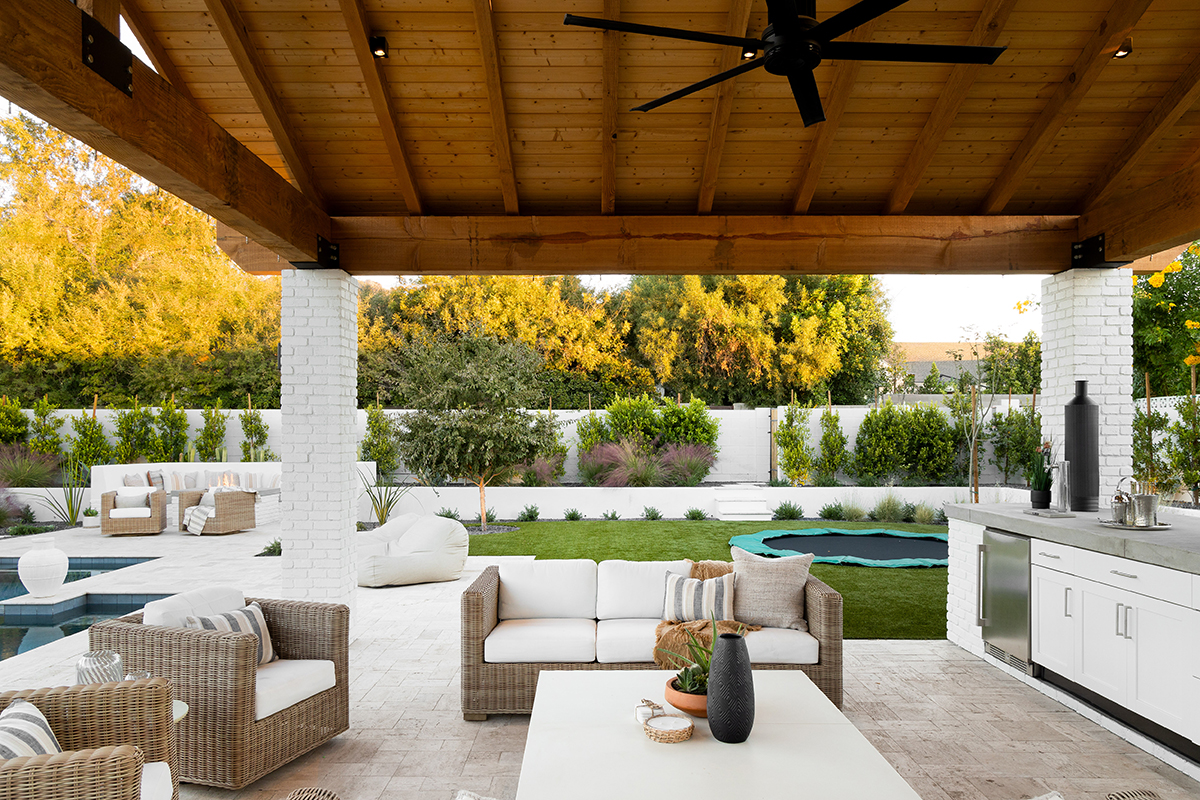 A beautiful and fun outdoor oasis perfect for family time and entertaining! We loved working with this Arcadia client to create the backyard of their dreams, which included a pool with a jump platform, a basketball / pickle ball court, and an in-ground trampoline! At the center of it all, the outdoor pavilion provides a central shaded space to kick back and relax whether that's watching the kids play in the pool or tuning into a game or movie while enjoying a brew from the kegerator built into the counter. Entertaining is a breeze with the custom bbq / kitchen space that features a raised counter with bar seating, and a fire pit with built-in seating. The plants and materials chosen represent the iconic Arcadia neighborhood style with a mixture of plantings such as oaks and dwarf olives, and classic finishes including travertine and white painted brick, while also exhibiting some fresh, transitionary elements such as grasses and cacti, concrete, and sharp, clean lines.  This Arcadia backyard exemplifies the seamless blend of indoor & outdoor spaces, a feature highly sought after by our clients. Pictured: View under ramada showing outdoor living room with view of in-ground trampoline in the background alongside the outdoor fire pit seating area. Backyard designed and built by On Site Landscape in Arcadia, Arizona.