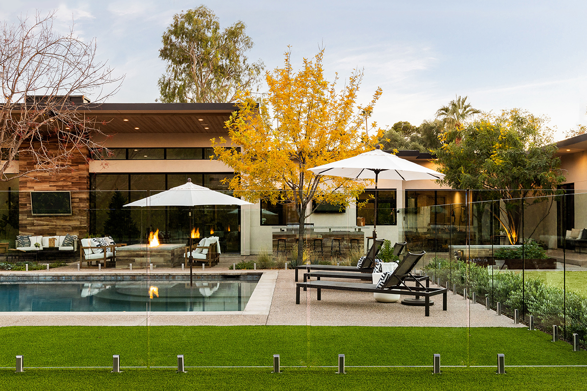 This landscape renovation project by On Site Landscape in Arcadia, Arizona, shows view of the backyard looking across the modern pool and spa with a glass pool enclosure, overlooking the turf lawn and outdoor living and dining areas