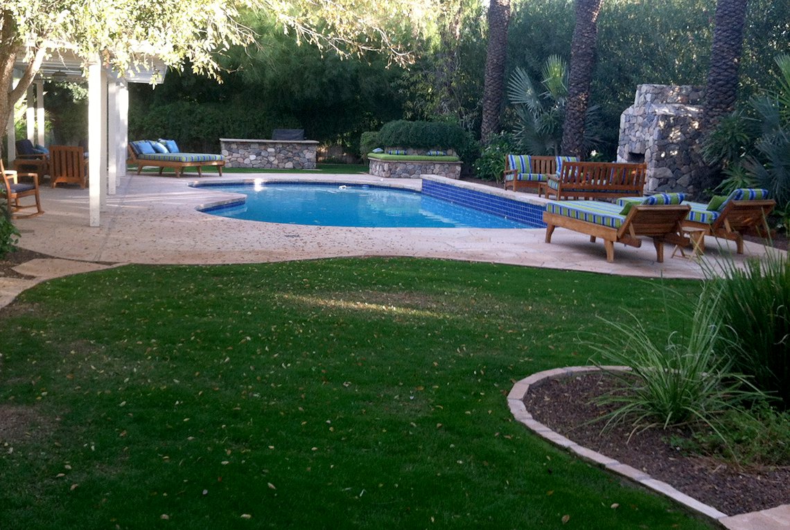 Tropical inspired landscape design by On Site Landscape in Paradise Valley, Arizona featuring a sparkling pool, covered patio, outdoor fireplace as well as lush plantings and a green lawn, perfect for family and friends spending time outside.